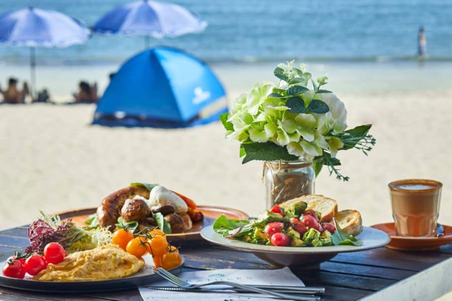 The breakfast at Bathers includes the Potato and Sinach Omelette, the Lantau Big Start (pork sausage, smoked bacon, grilled tomato, mushrooms, poached egg & toasted sourdough), and Toasted Sourdough with Fresh Avocado. These dishes are on a wooden table that overlooks bathers and tents pitched on the beach.