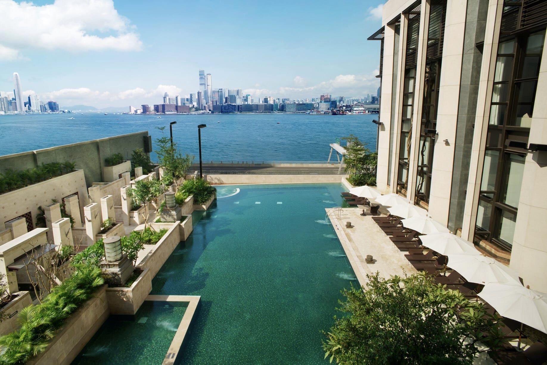 The swimming pool at the Harbour Grand Hong Kong has a children's pool and landscaping on the left, and deck chairs with individual umbrellas on the right. It overlooks Kowloon and has views of Hong Kong Island.