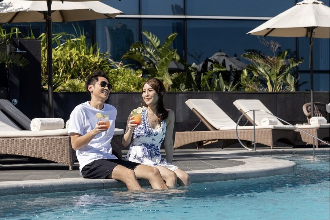A couple sit by the pool of the Kerry Hotel while their legs dangle in the water. The man on the left is wearing a white T-shirt and black shorts and the woman on the right is wearing a white sundress with blue flowers. They're both holding a glass with a cocktail. Behind them are deck chairs, umbrella and the hotel landscaping.