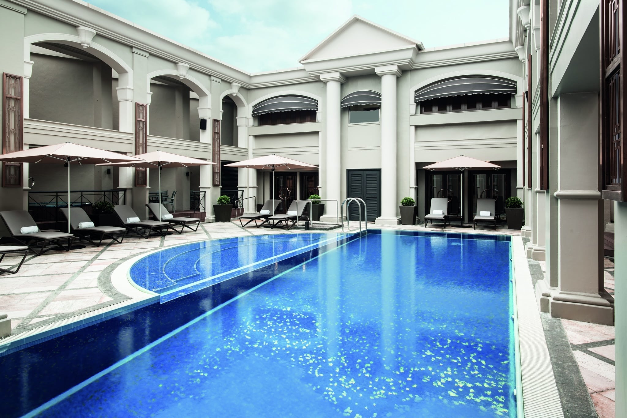 The swimming pool at The Langham Hong Kong surrounded by lounge chaises sheltered by umbrellas. There are columned corridors all around the pool.