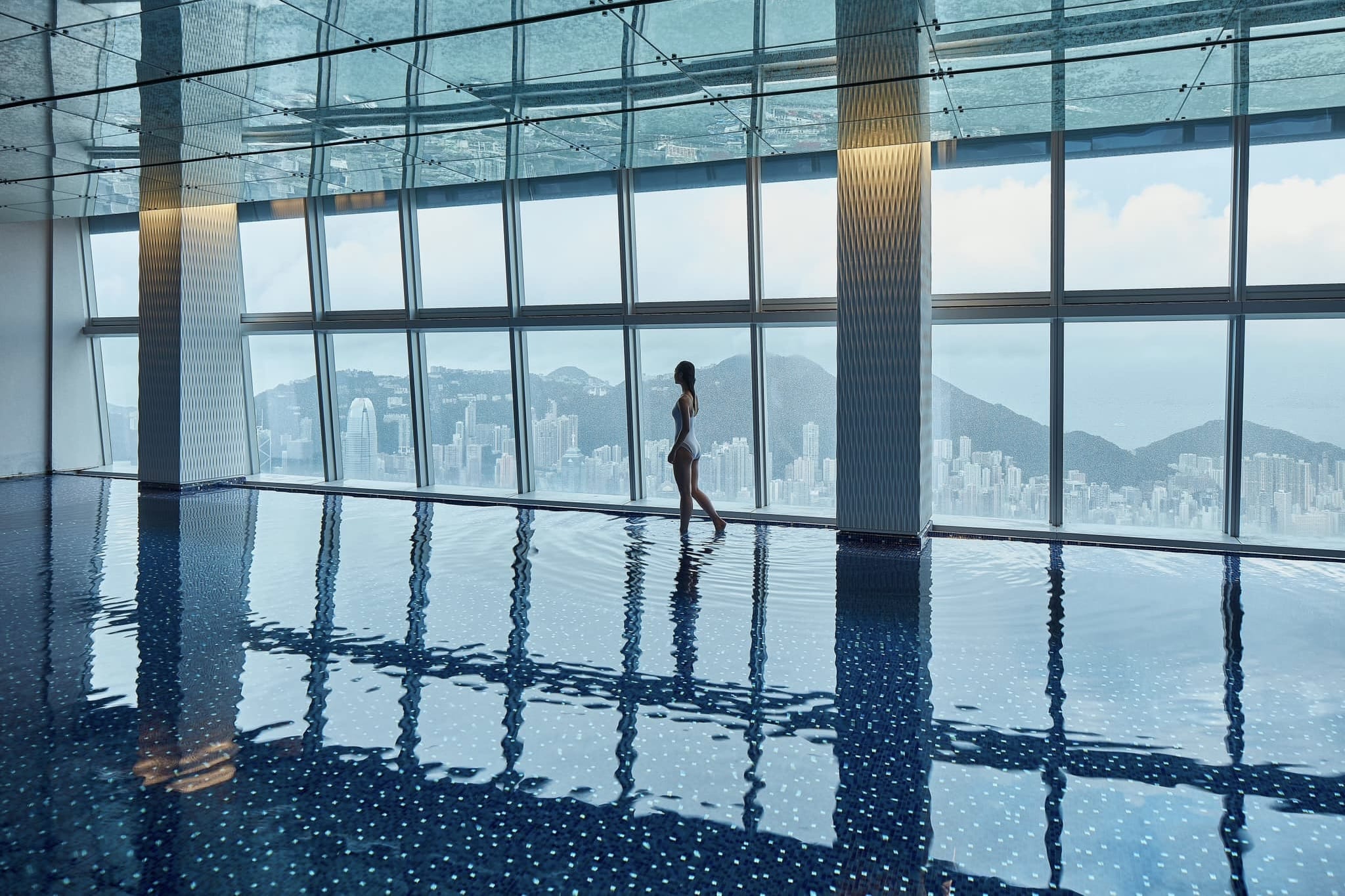 A woman in a swimsuit walks in the pool at The Ritz-Carlton Hong Kong. She walks in front of the floor-to-ceiling windows of the pool, which overlooks Hong Kong Island.
